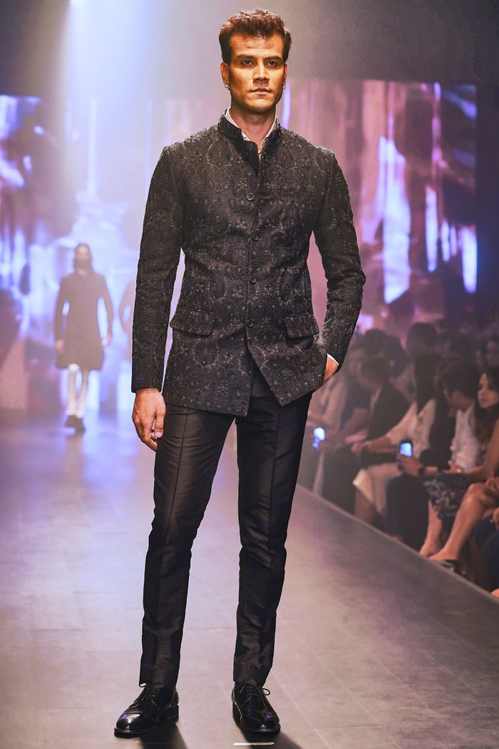 Male model in a black outfit walks the runway during a fashion show, showcasing modern style.