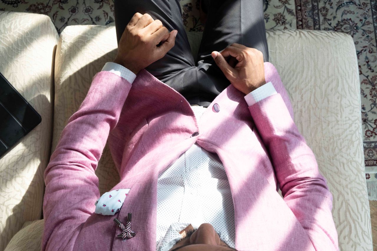 A man in a pink blazer relaxes on a sofa in a sunlit Dubai apartment.