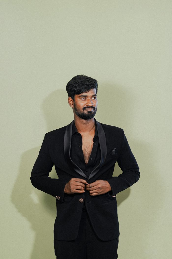 Stylish man wearing a black suit poses confidently against a simple pale green background.
