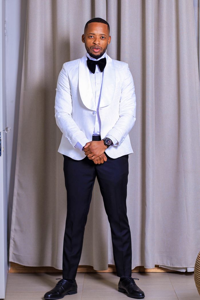 A stylish man wearing a white tuxedo and bow tie, posing indoors against curtains.