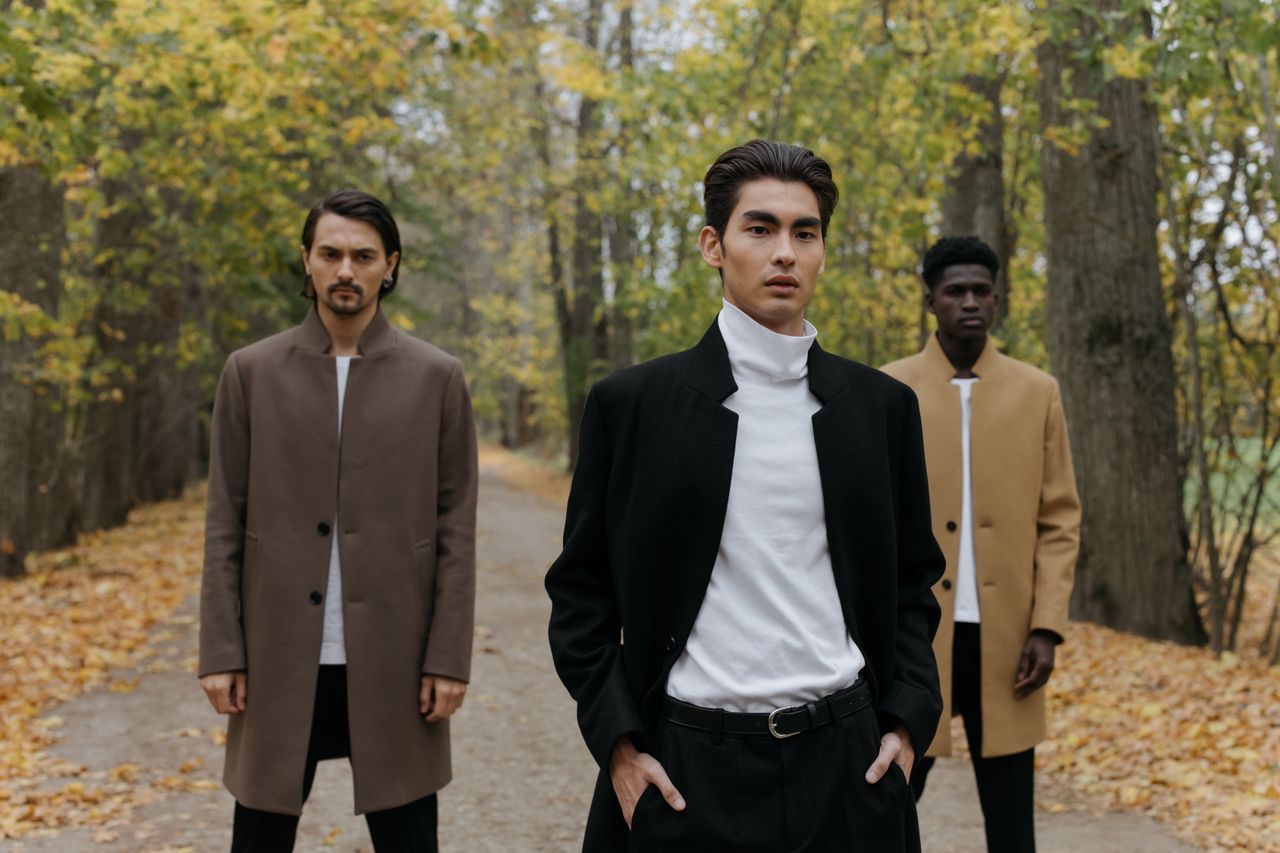 Three men in stylish coats standing on a leafy path in autumn.